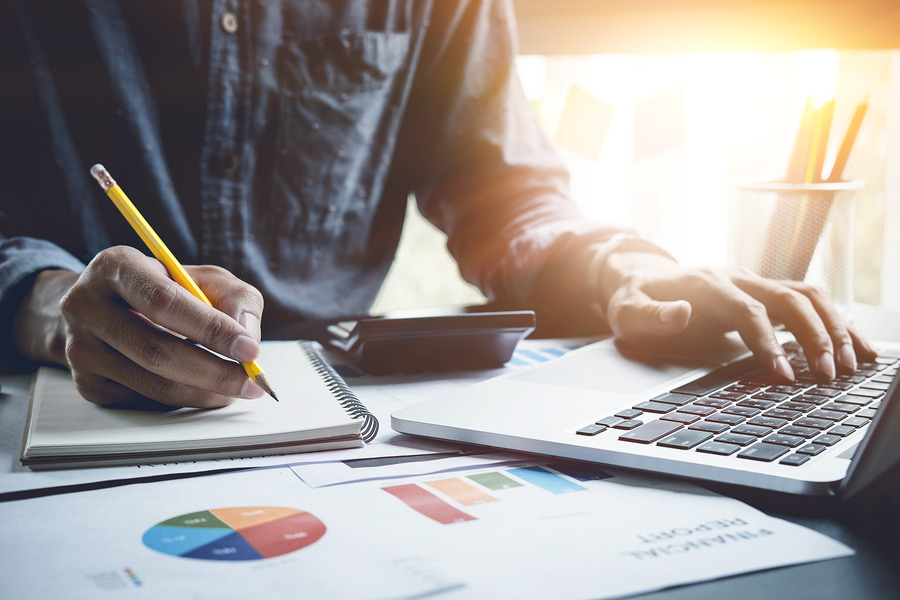 Man taking notes beside a laptop and financial charts