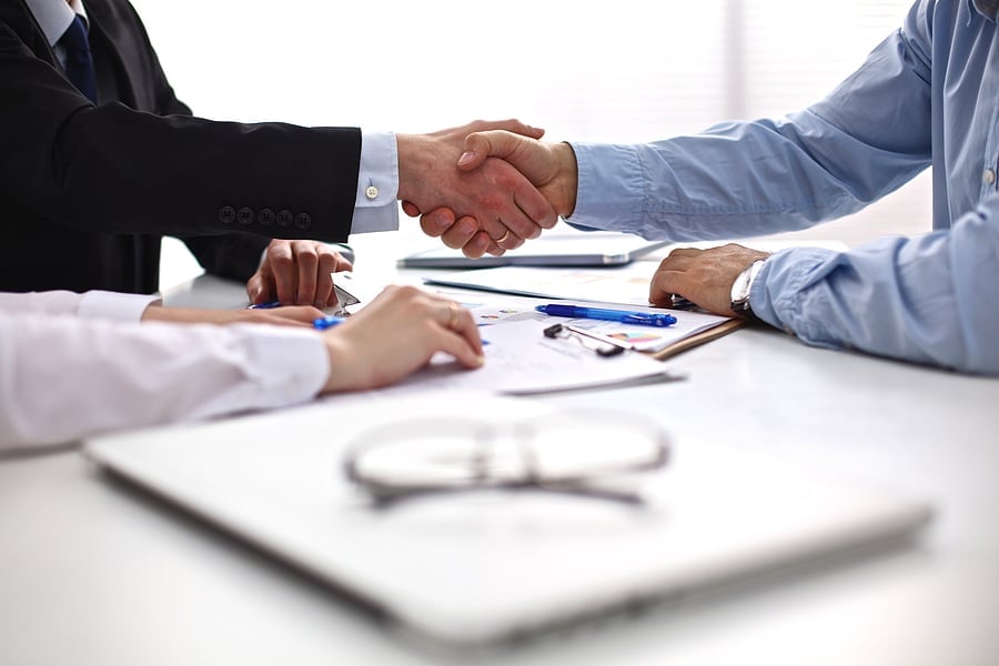 Two colleagues shaking hands across a table with paperwork and coffee