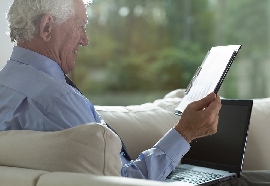 Retired man reviewing documents on a sofa with a laptop nearby